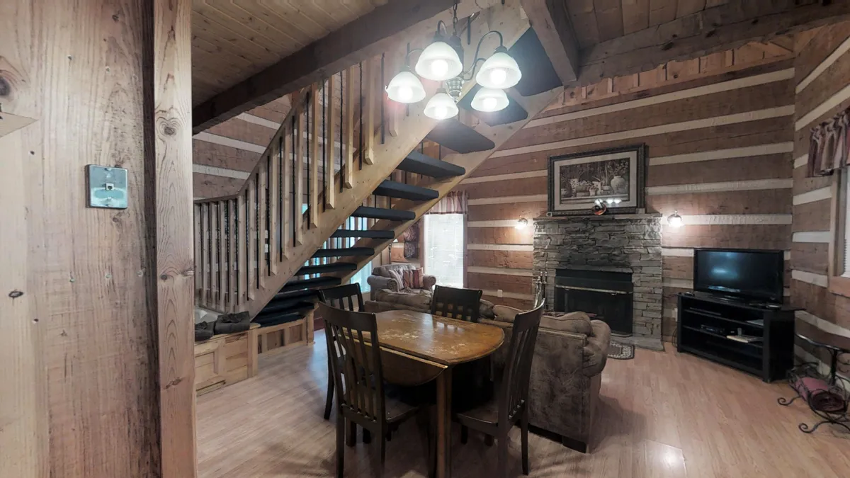 A warm, inviting interior featuring a rustic stone fireplace surrounded by cozy seating, a wooden staircase, and a dining table. The space exudes comfort, making it ideal for intimate evenings spent together.