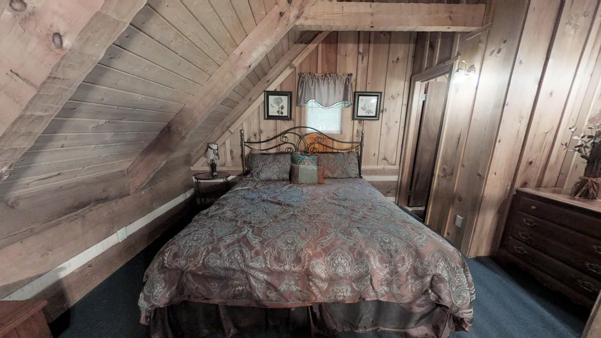 A cozy loft bedroom featuring a king-size bed nestled beneath rustic wooden beams, with a dressing table and framed pictures on the wall. Warm lighting and inviting decor create an intimate, comfortable atmosphere perfect for couples.