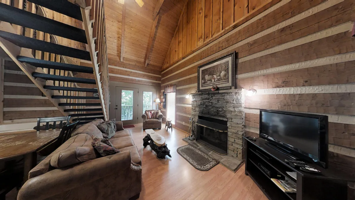 A warm and welcoming living room with a stone fireplace, wooden beams, a comfortable seating area, and large windows letting in natural light. The arrangement is open and spacious, capturing the inviting atmosphere described in the section. The visible fireplace and seating area evoke coziness and togetherness, ideal for relaxing evenings.
