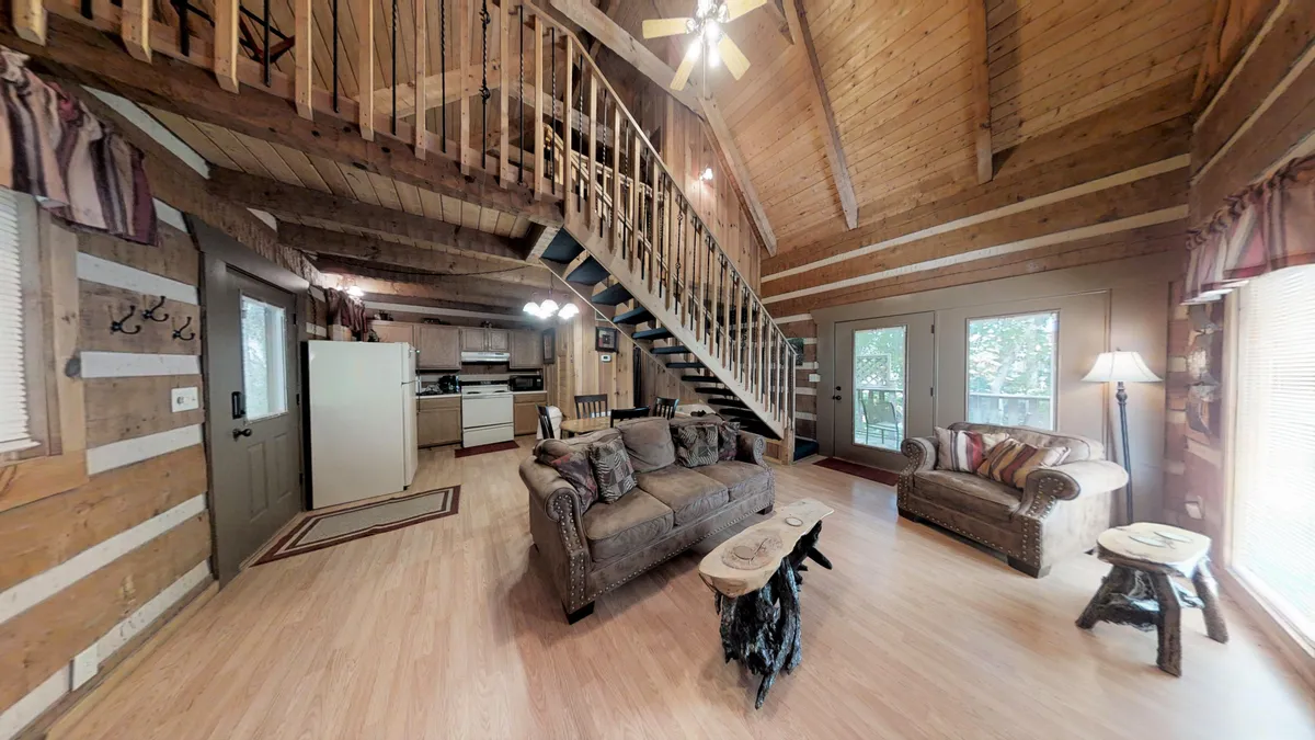This inviting interior scene shows a spacious area that merges the kitchen, a comfortable seating area, and a dining table. The kitchen area features cabinetry and modern appliances, while the dining table is set up for shared meals. Large windows usher in natural light, creating a warm and welcoming environment.