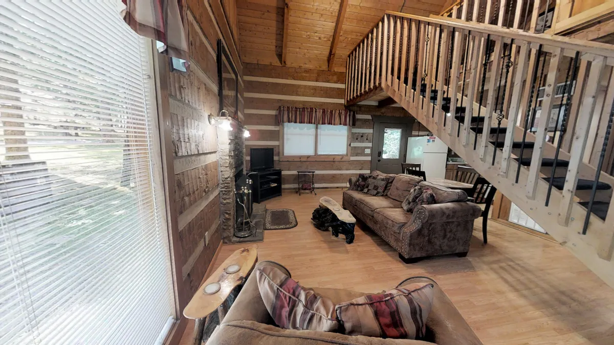 An inviting living room scene showing a comfortable sofa, a stone fireplace, a staircase, and large windows. The setup is cozy yet open, fostering togetherness with ample seating and a view of the crackling fire, ideal for quiet nights in or socializing with loved ones.
