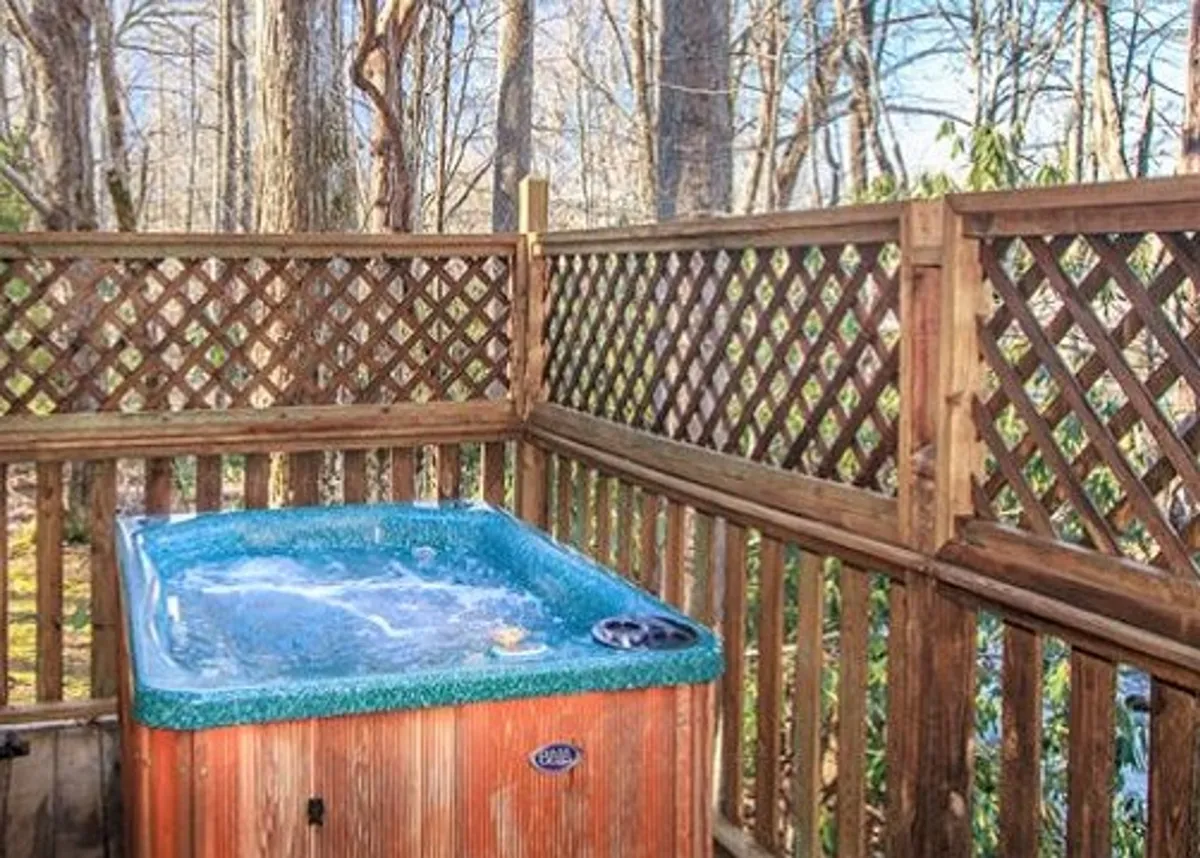 This exterior image highlights a spacious hot tub nestled on a wooden deck, surrounded by trees and a rustic wooden fence. The tub is positioned for privacy while still offering open views of the natural surroundings, with sunlight filtering through the foliage and a tranquil outdoor mood.